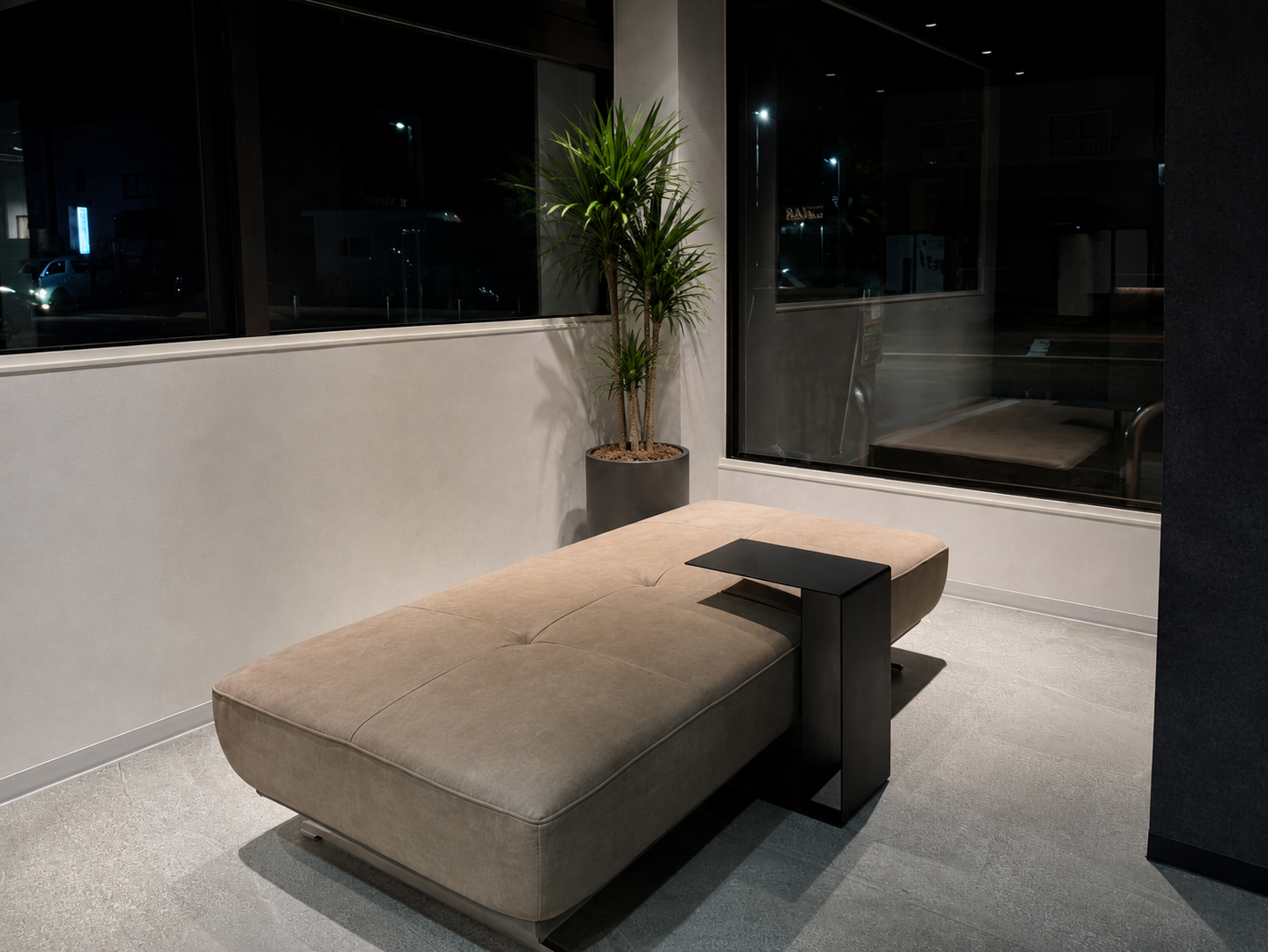 Minimalist hotel lobby seating: beige chaise longue with a black metal side table, next to a tall potted plant by a window at night