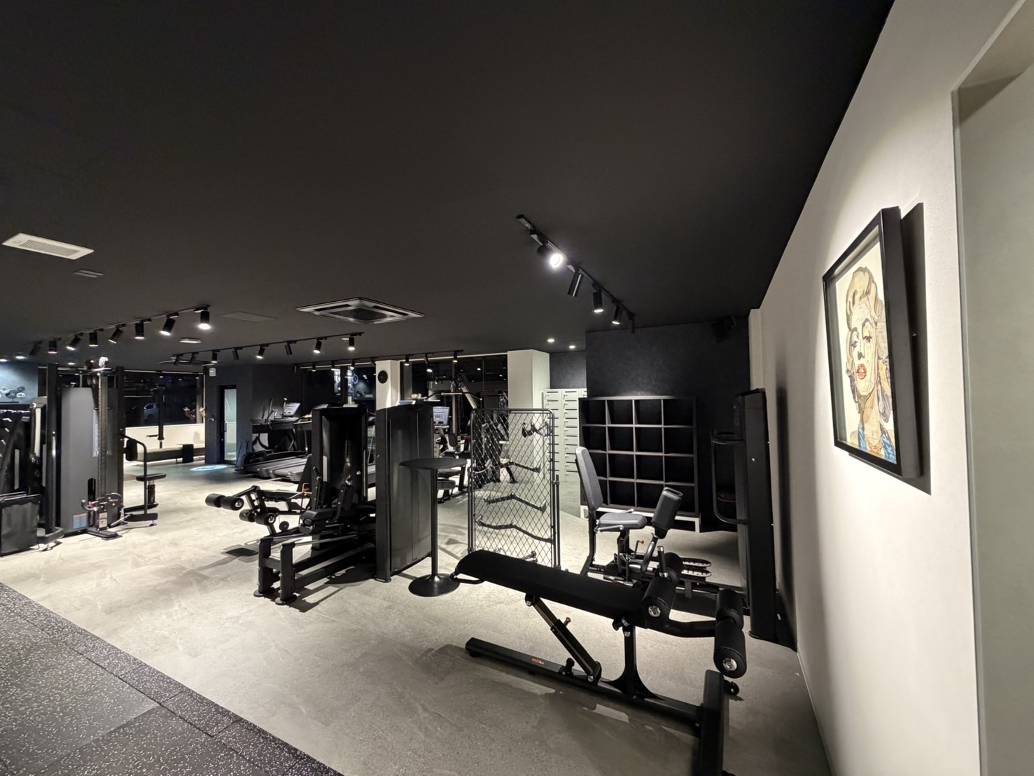 Well‑lit indoor gym with black ceiling and gray flooring, featuring weight machines, benches, and a metal partition in the center.