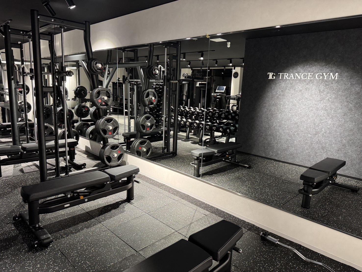 Strength training area with racks of barbells, plates, and benches, reflected in a large mirror on the wall.