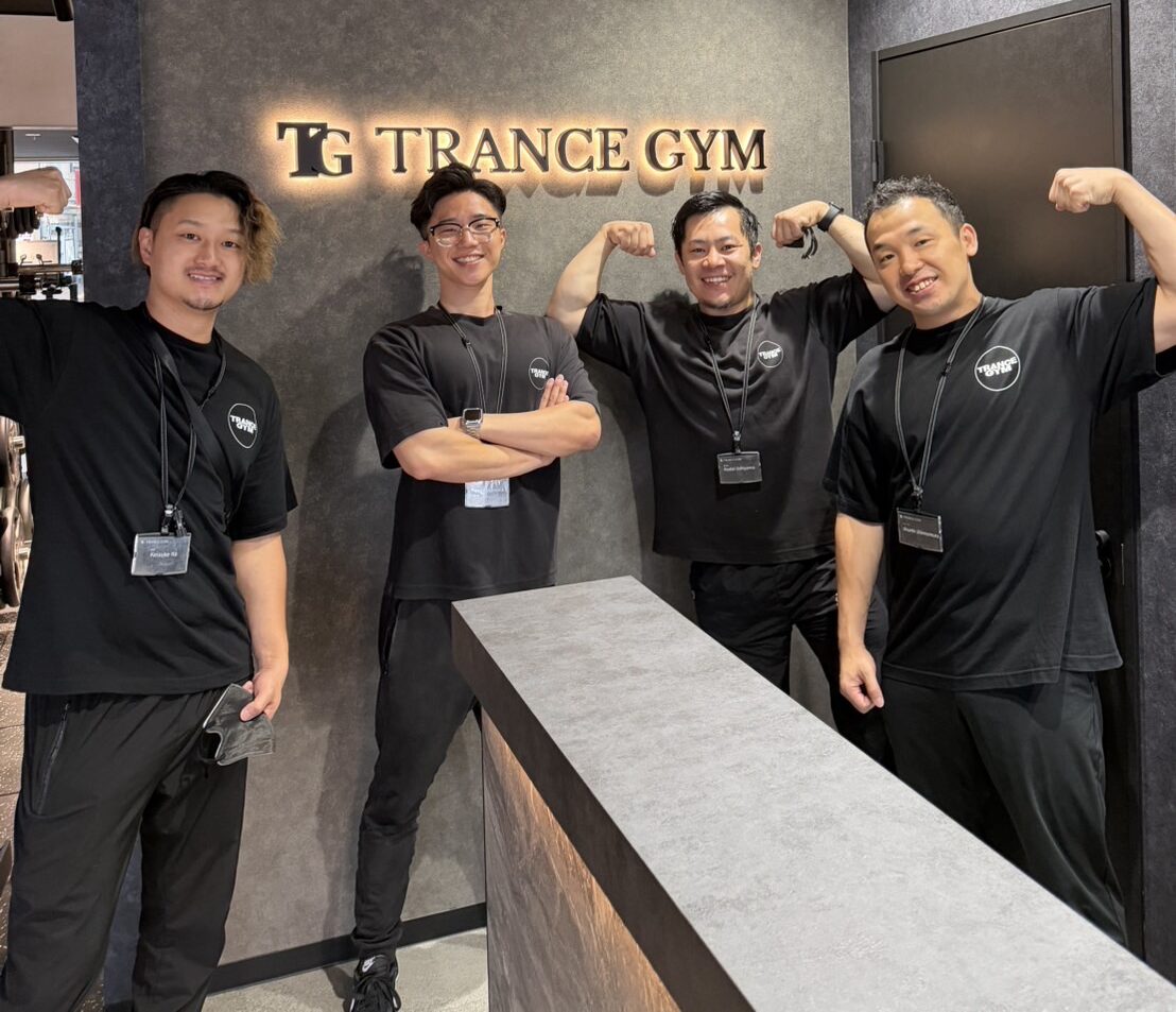 Four staff members in black uniforms pose and flex their arms in front of a TG Trance Gym sign on a dark wall in the lobby area.