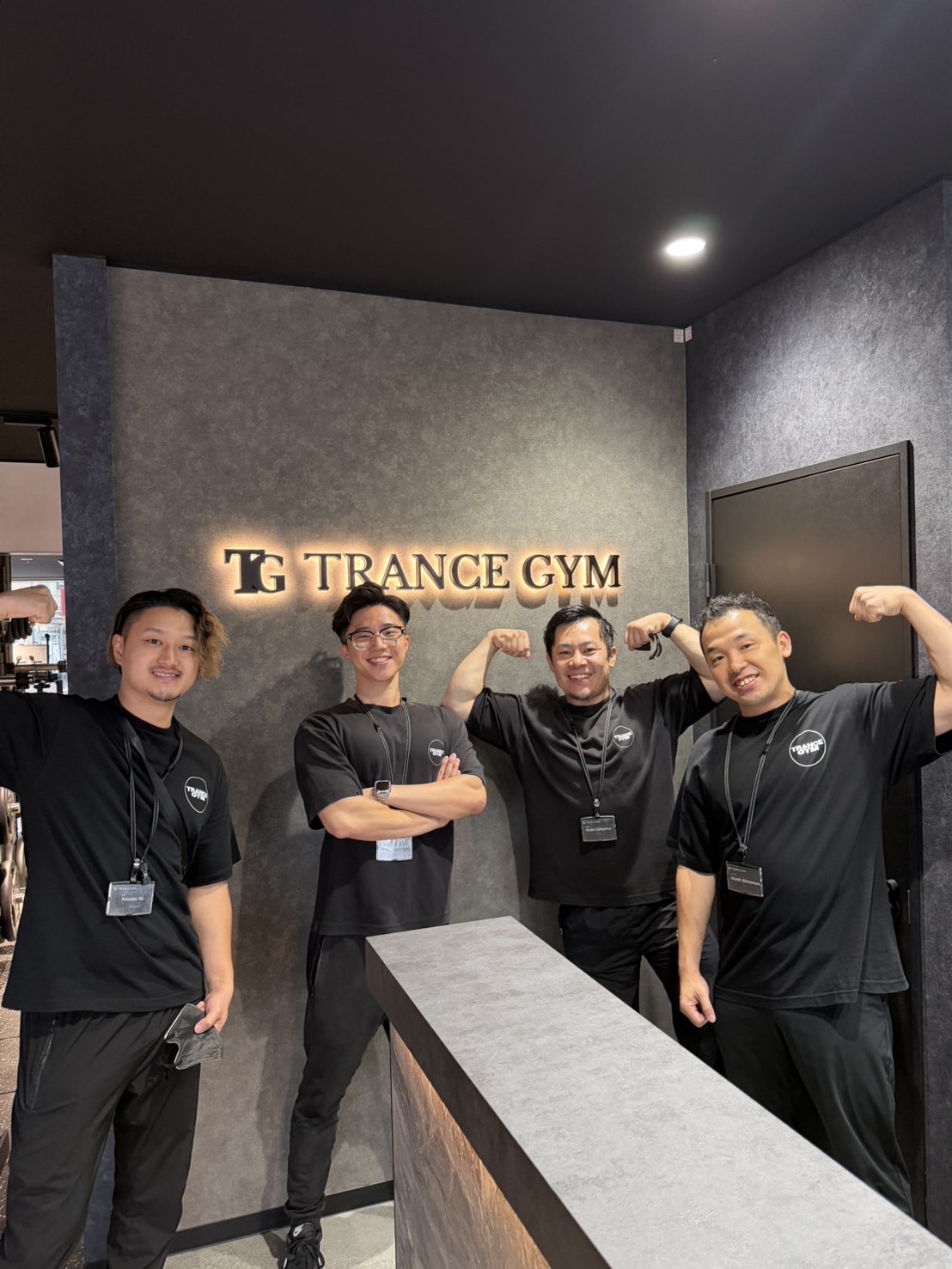 Four staff members in black uniforms pose and flex their arms in front of a TG Trance Gym sign on a dark wall in the lobby area.